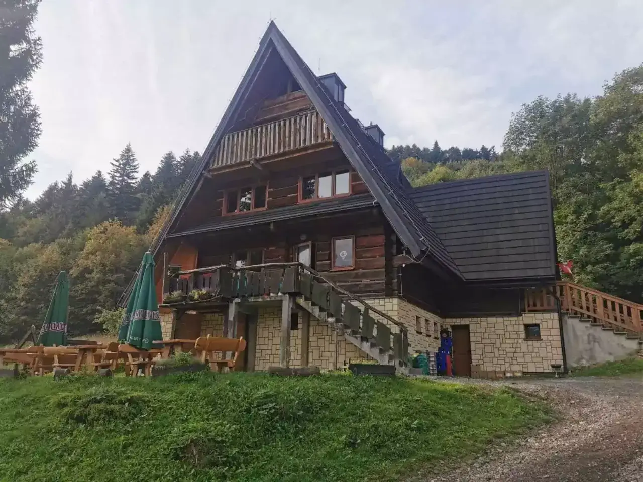 Drewniana bacówka pod honem, otoczona lasem, z tarasem i stolikami na zewnątrz.